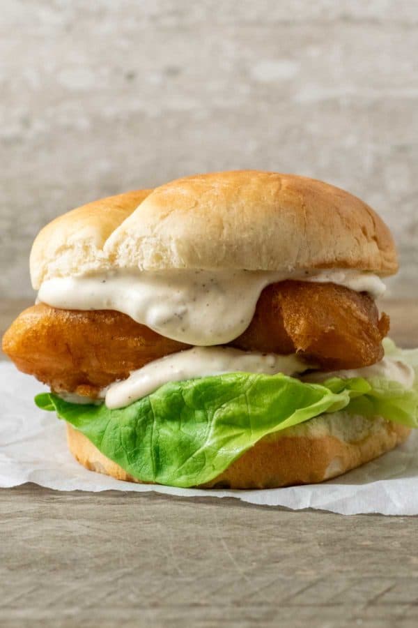 a bun filled with lettuce, white creamy sauce, and golden brown fish filet on parchment paper.
