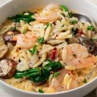 a bowl filled with shrimp, orzo pasta, mushrooms, spinach, and roasted red peppers in a yellow creamy sauce and a spoon scooping some out