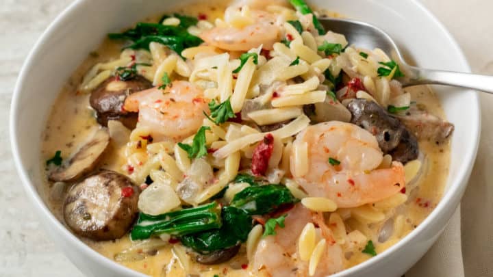 a bowl filled with shrimp, orzo pasta, mushrooms, spinach, and roasted red peppers in a yellow creamy sauce and a spoon scooping some out