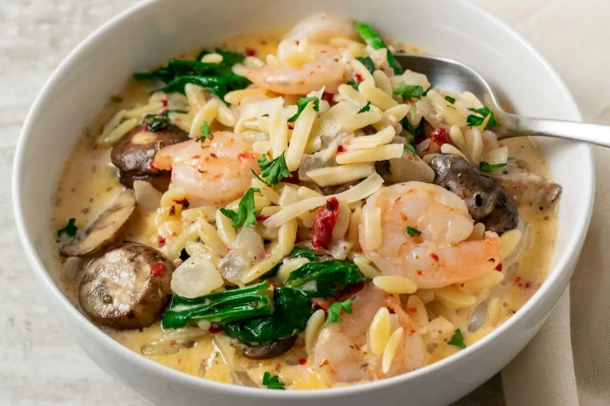 a bowl filled with shrimp, orzo pasta, mushrooms, spinach, and roasted red peppers in a yellow creamy sauce and a spoon scooping some out