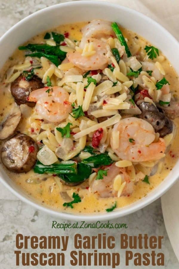 a bowl filled with shrimp, orzo pasta, mushrooms, spinach, and roasted red peppers in a yellow creamy sauce and text saying creamy garlic butter tuscan shrimp pasta recipe at zonacooks.com