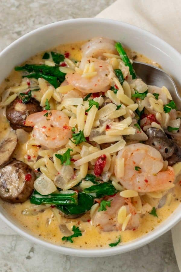 a bowl with a spoon scooing out some shrimp, orzo pasta, mushrooms, spinach, and roasted red peppers in a yellow creamy sauce
