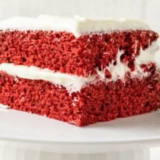 a white cake stand with a piece of two layer red cake with white creamy frosting