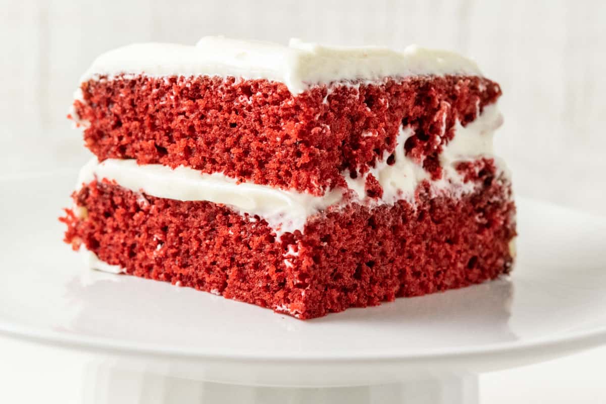a white cake stand with a piece of two layer red cake with white creamy frosting