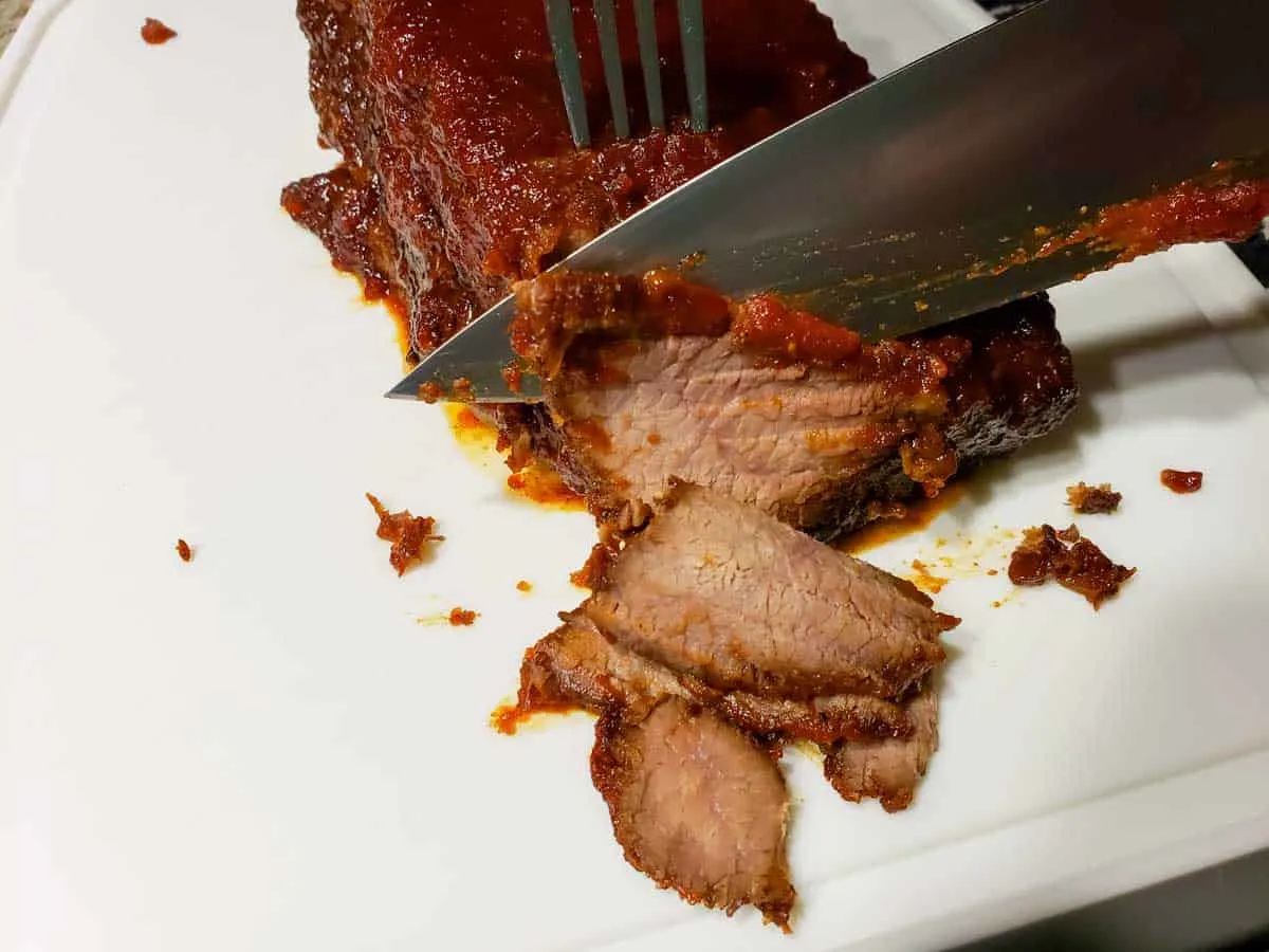 a fork holding a bbq brisket in place and a chef knife cutting thin slices off the end of it