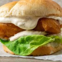 a bun filled with lettuce, white creamy sauce, and golden brown fish filet on parchment paper.