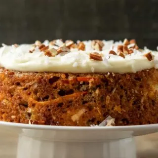 a mini carrot cake topped with cream cheese, coconut, and pecans on a cake stand in the background