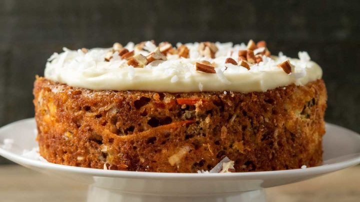 a mini carrot cake topped with cream cheese, coconut, and pecans on a cake stand in the background