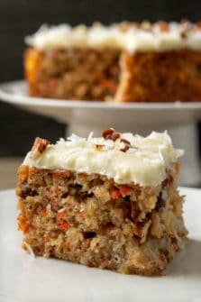 a slice of cake filled with carrots and pineapple and a small batch carrot cake topped with cream cheese on a cake stand in the background