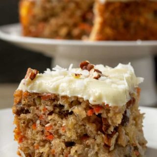 a slice of cake filled with carrots and pineapple and a small batch carrot cake topped with cream cheese on a cake stand in the background