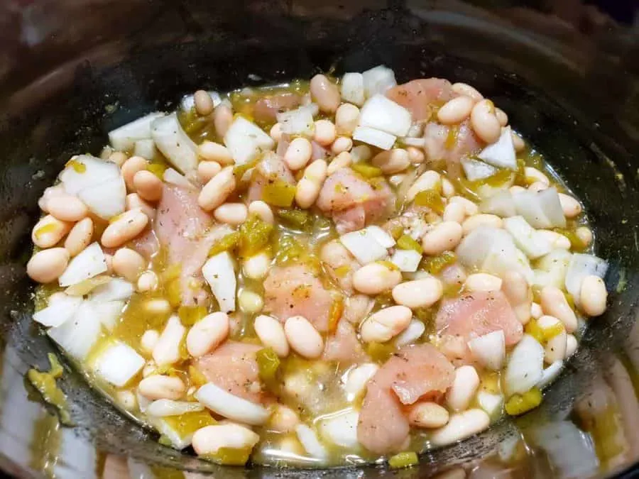 a slow cooker crock pot filled with diced onions, diced raw chicken, seasonings, northern beans, and clear broth