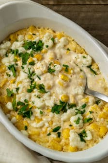 a casserole dish filled with corn, white cheese, chopped cilantro and creamy sauce with a spoon lifting some out