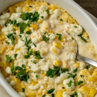 a casserole dish filled with corn, white cheese, chopped cilantro and creamy sauce with a spoon lifting some out