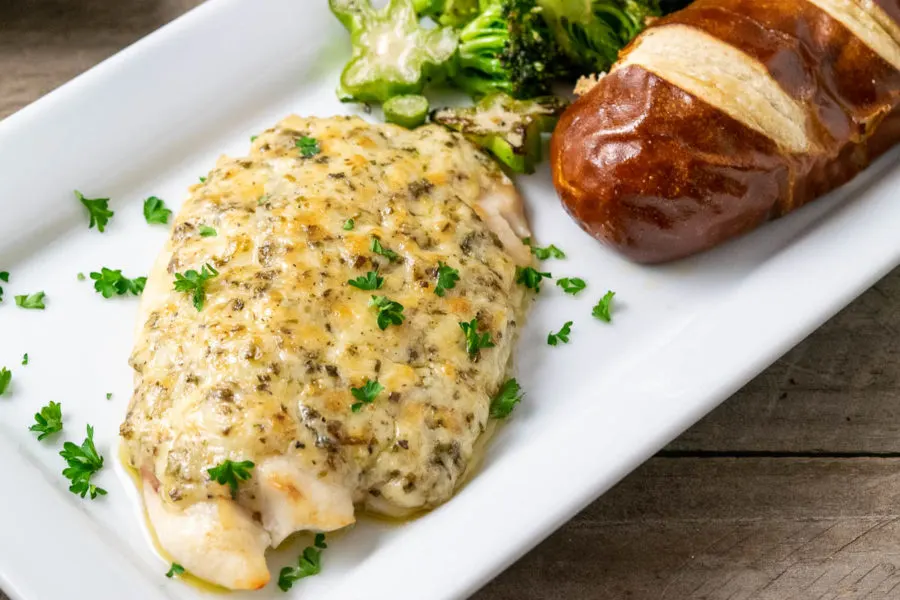 a plate filled with a piece of Tilapia fish topped with a Parmesan and mayo mixture and sides of broccoli and pretzel baguette