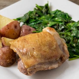 a close up front view of a golden brown chicken thigh with sides of red potatoes and spinach coated in sauce