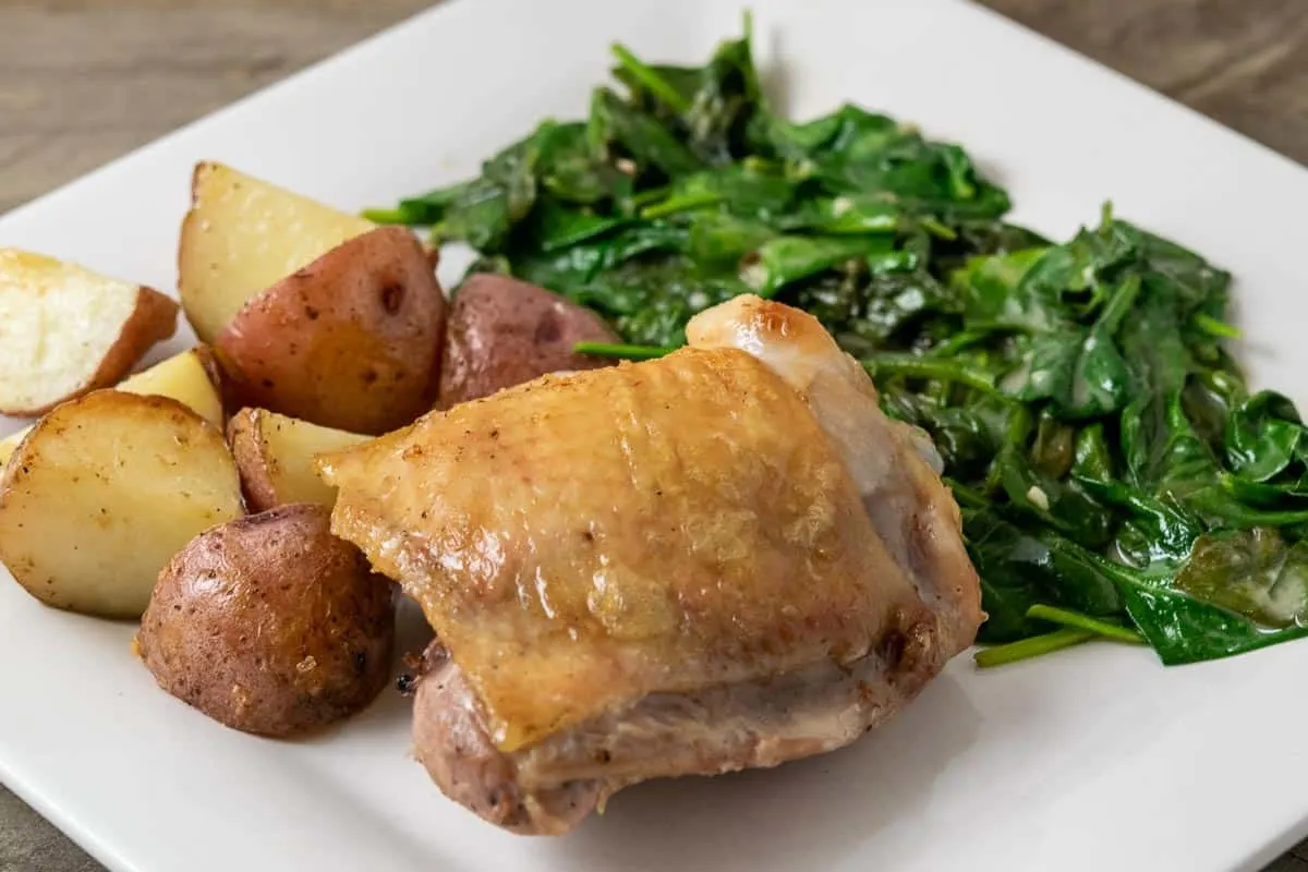 a close up front view of a golden brown chicken thigh with sides of red potatoes and spinach coated in sauce