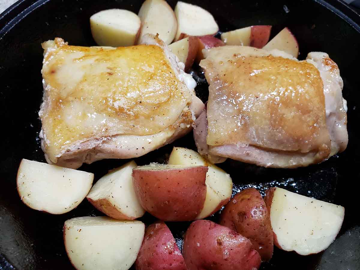 two browned chicken thighs red potatoes cooking in a cast iron skillet