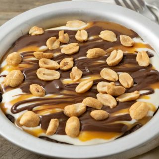 a small pie pan filled with chocolate crust, creamy filling, caramel sauce drizzle, chocolate ganache drizzle, and peanuts and two forks.