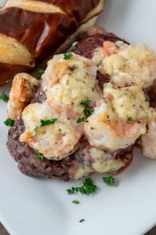 a filet mignon steak topped with shrimp and Parmesan cheese sauce sprinkled with parsley and a side of pretzel baguette