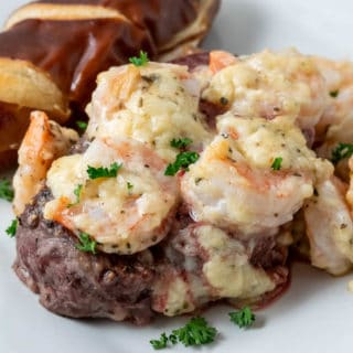 a close up of a plate filled with a filet mignon steak topped with shrimp and Parmesan cheese sauce with a side of pretzel baguette