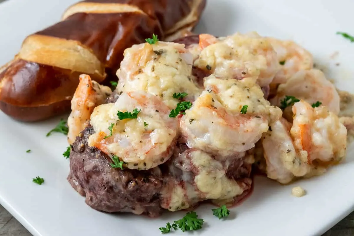 a close up of a plate filled with a filet mignon steak topped with shrimp and Parmesan cheese sauce with a side of pretzel baguette