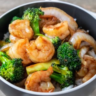 a close up of a bowl filled with white rice, broccoli florets, sliced onions, and shrimp coated in an orange colored sauce