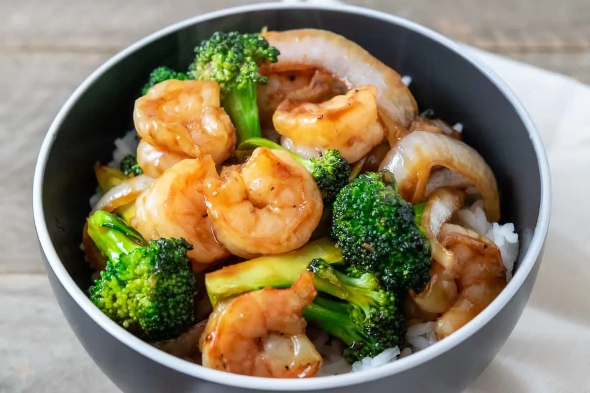 a close up of a bowl filled with white rice, broccoli florets, sliced onions, and shrimp coated in an orange colored sauce.