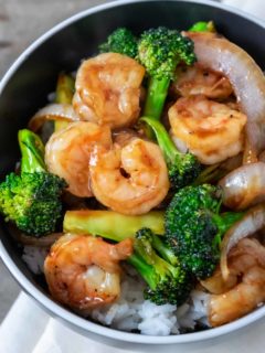 a black bowl filled with white rice, broccoli florets, sliced onions, and shrimp coated in an orange colored sauce