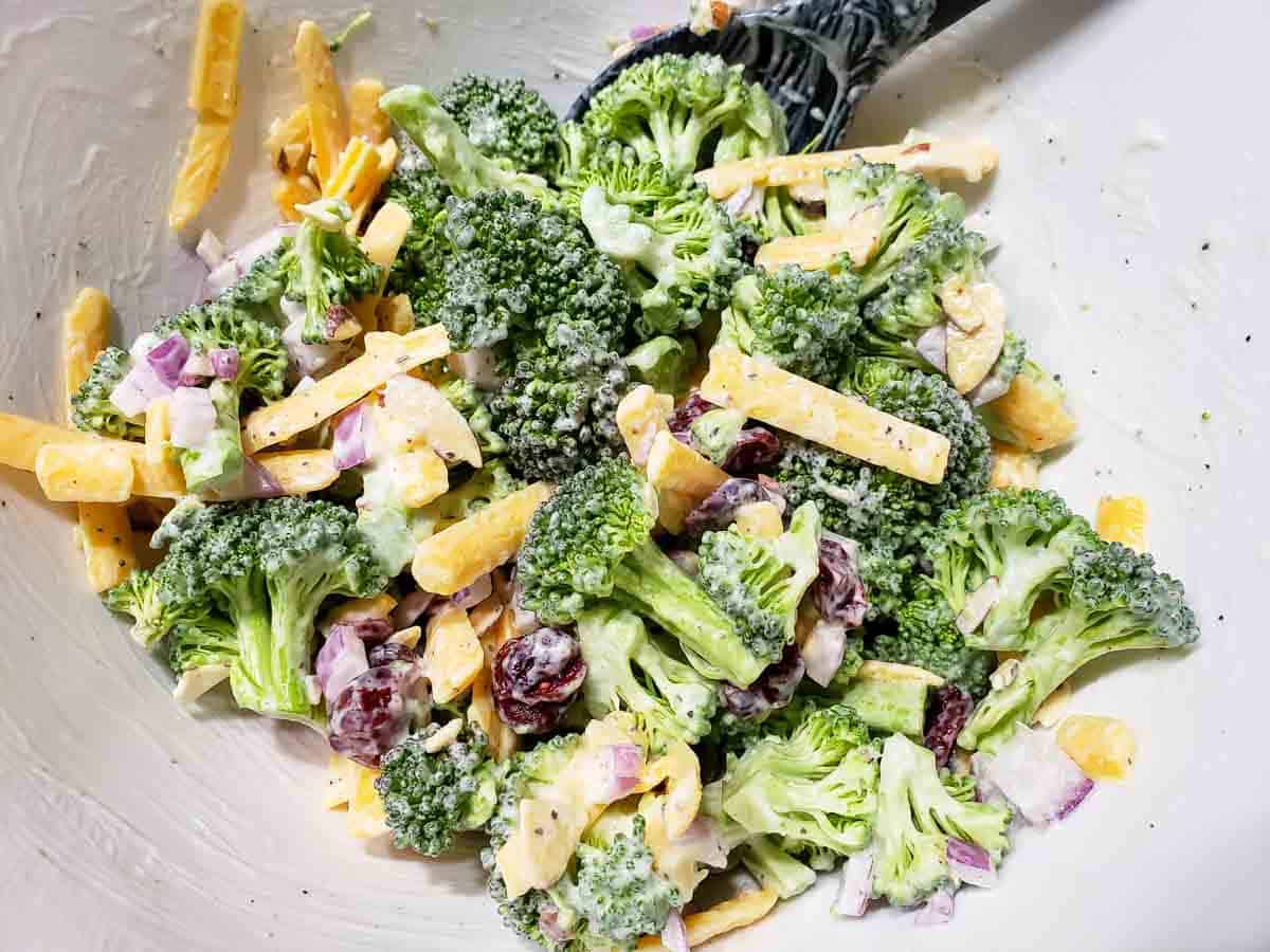 broccoli florets, diced red onion, dried cranberries, and shredded cheddar cheese coated in white creamy mayo in a bowl with a black spoon
