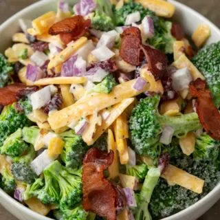 a close up front view of a bowl filled with broccoli, red onion, bacon, cranberries and shredded cheese coated in a white creamy sauce