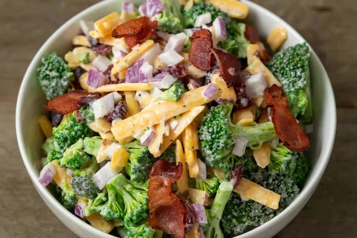 a close up front view of cold broccoli salad with a bowl filled with broccoli, red onion, bacon, cranberries and shredded cheese coated in a white creamy sauce