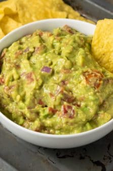 a bowl of mashed green avocado and tomato mixture and a side of tortilla chips and a single chip dipped in the guac