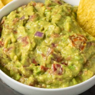 a bowl of mashed green avocado and tomato mixture and a side of tortilla chips and a single chip dipped in the guac