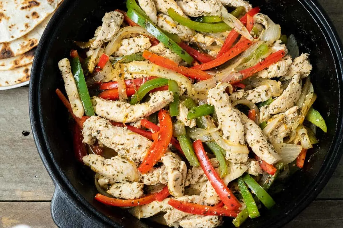 a cast iron skillet filled with sliced green pepper, red pepper, onions, and chicken in homemade fajita seasoning and fire roasted tortillas.