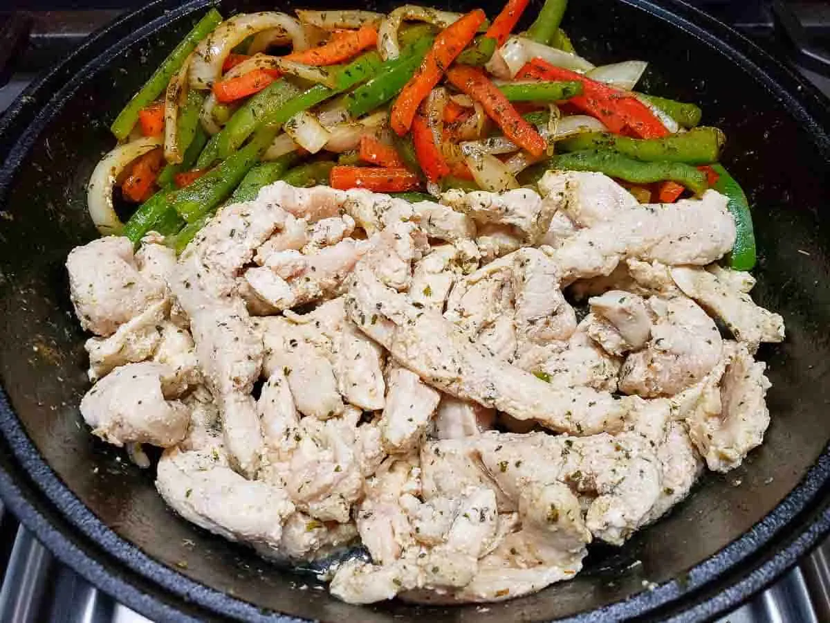 sliced green peppers, red peppers and onions coated in the best fajita seasoning next to boneless chicken slices cooking in a cast iron skillet.