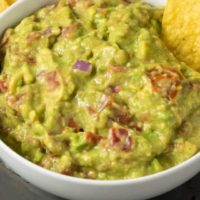 a bowl of mashed green avocado and tomato mixture and a side of tortilla chips and a single chip dipped in the guac