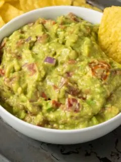 a bowl of mashed green avocado and tomato mixture and a side of tortilla chips and a single chip dipped in the guac