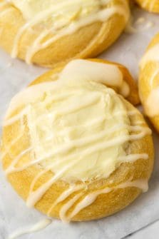 a close up of golden brown crescent dough circles topped with a cream cheese filling in the center and a white creamy glaze drizzle