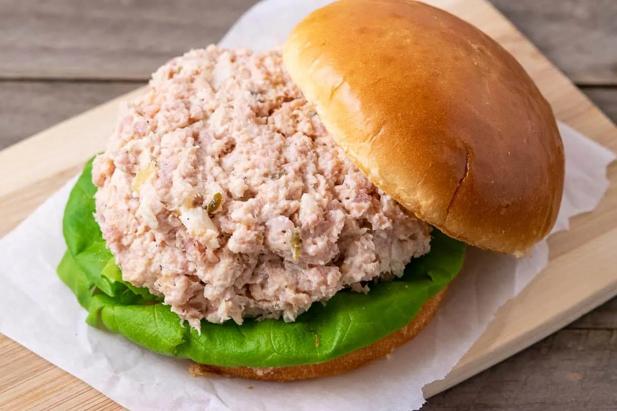 a sandwich roll bottom filled with lettuce and ham salad spread and the sandwich roll bun top resting on the edge of it