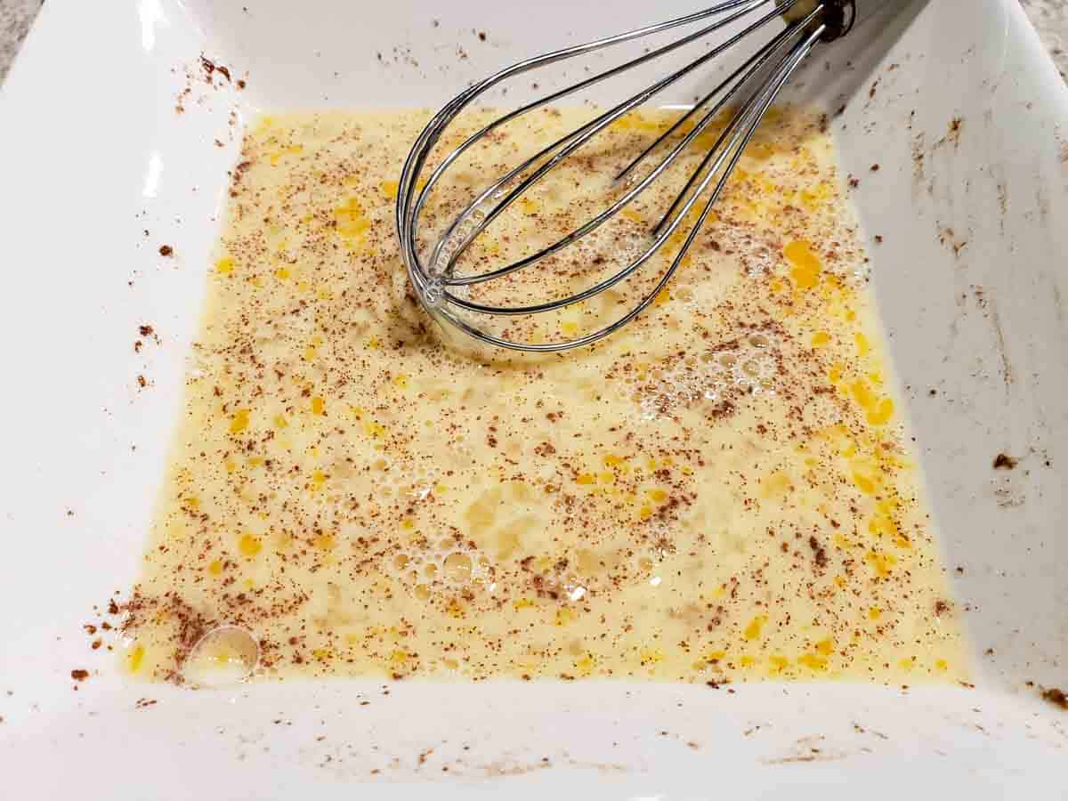 a square bowl filled with uncooked whisked egg, milk, vanilla, sugar, cinnamon, and salt mixture.