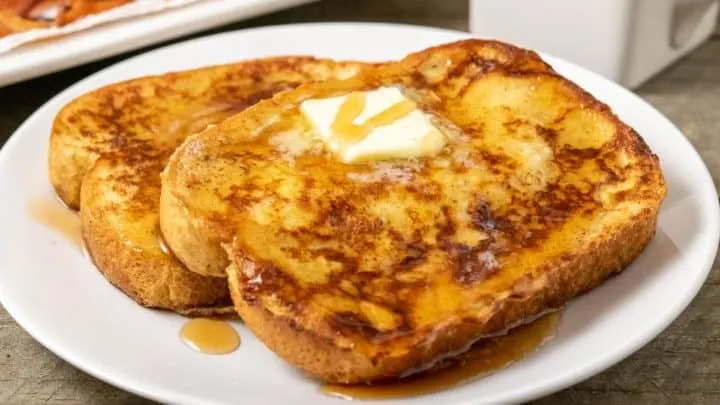 two thick slices of texas toast french toast topped with butter and syrup with a side of bacon and milk.