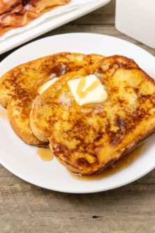 two thick slices of texas toast french toast topped with butter and syrup and a side of bacon.