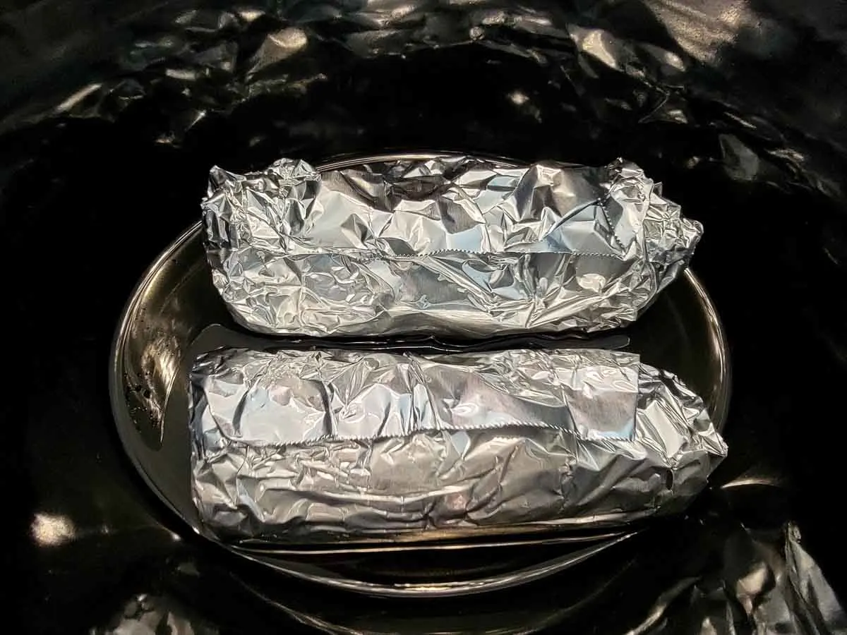 two corn on the cobs wrapped in tinfoil sitting in a crock pot.