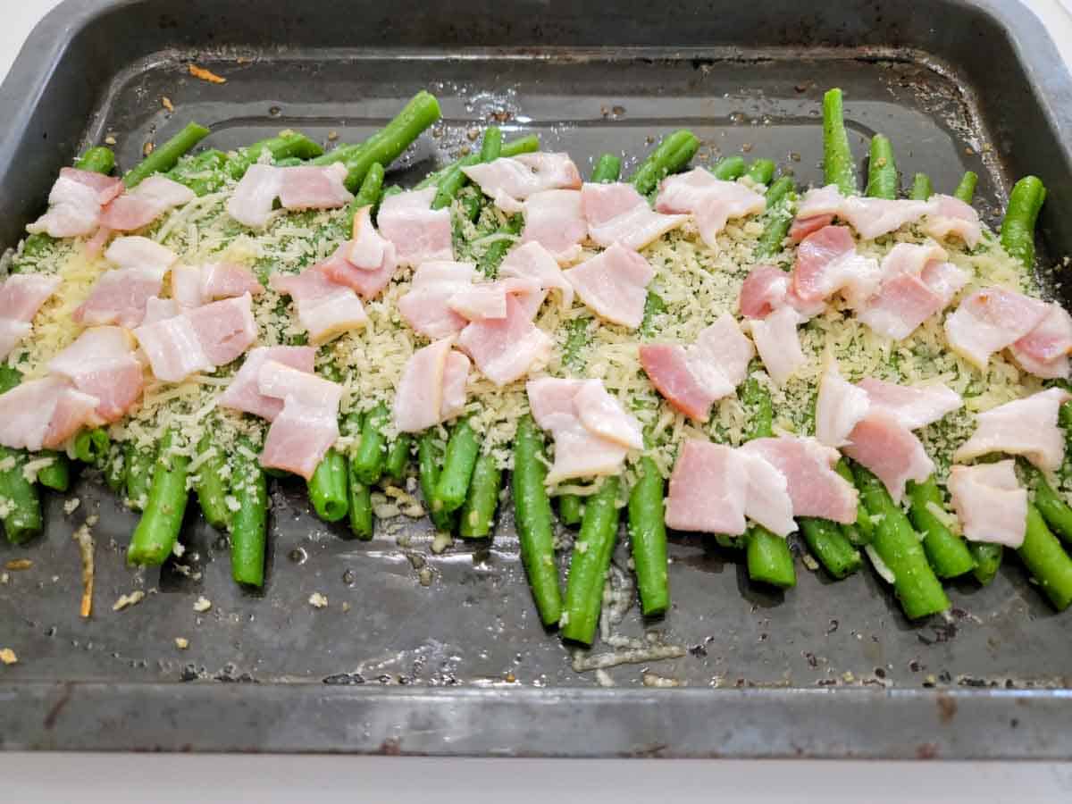green beans in a row topped with panko bread crumbs, parmesan cheese, and bacon pieces on a baking sheet.