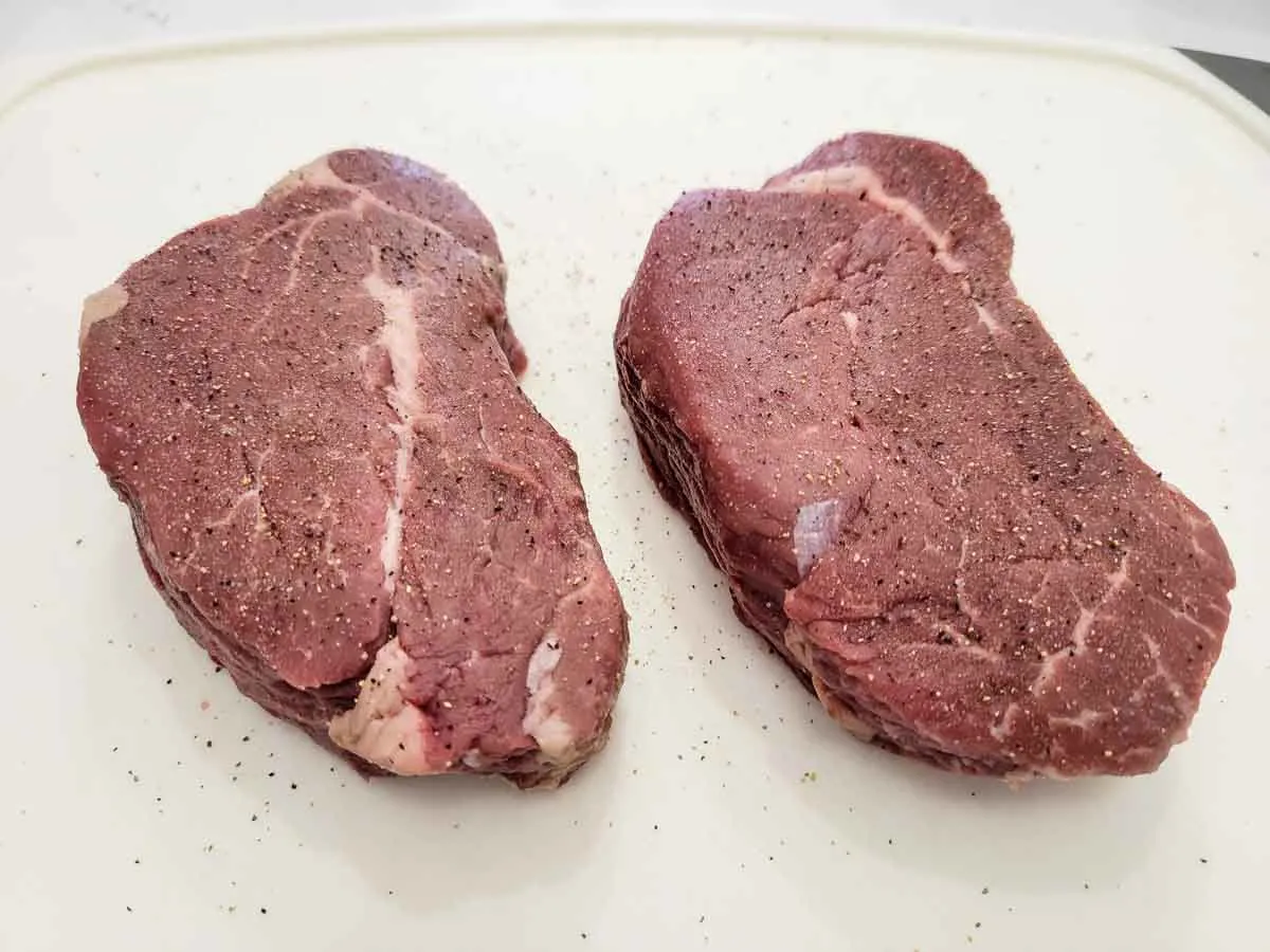 two filet mignon medallions sprinkled with salt and pepper on a cutting board.