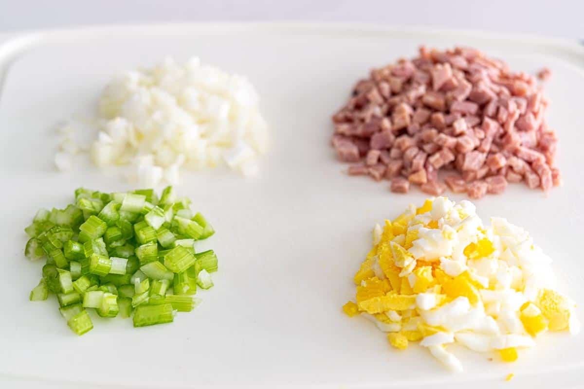 a cutting board with piles of diced onions, ham, celery, and hard-boiled egg.