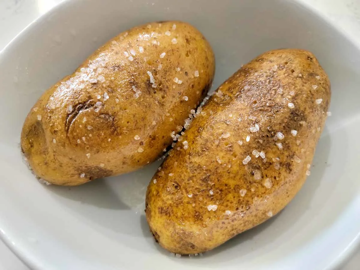 two russet potatoes coated in oil and coarse salt in a bowl.