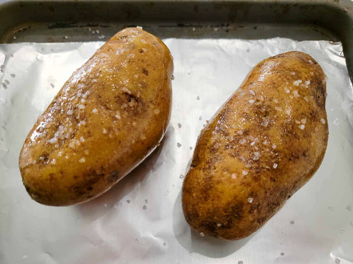 two russet potatoes coated in oil and coarse salt on a foil lined baking sheet.