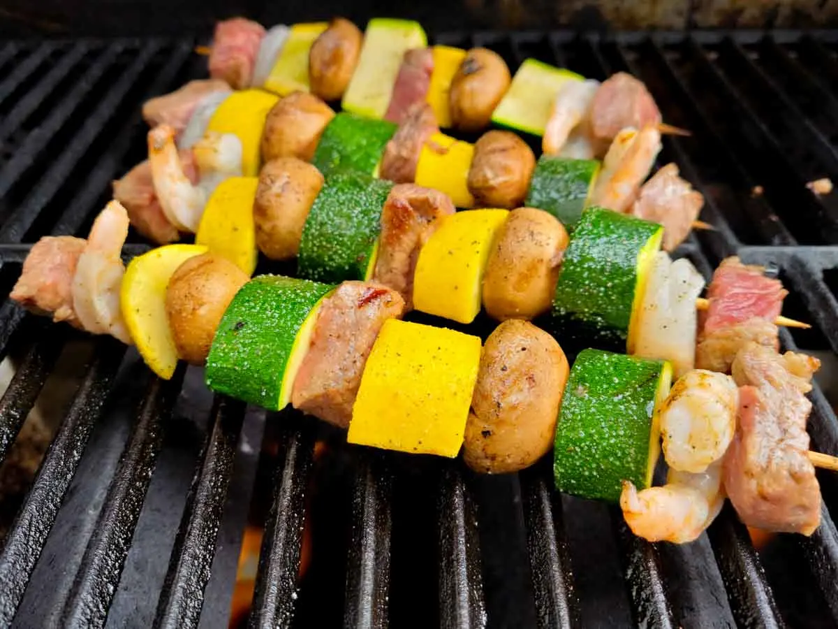 beef, shrimp, and veggie kabobs cooking on a grill.