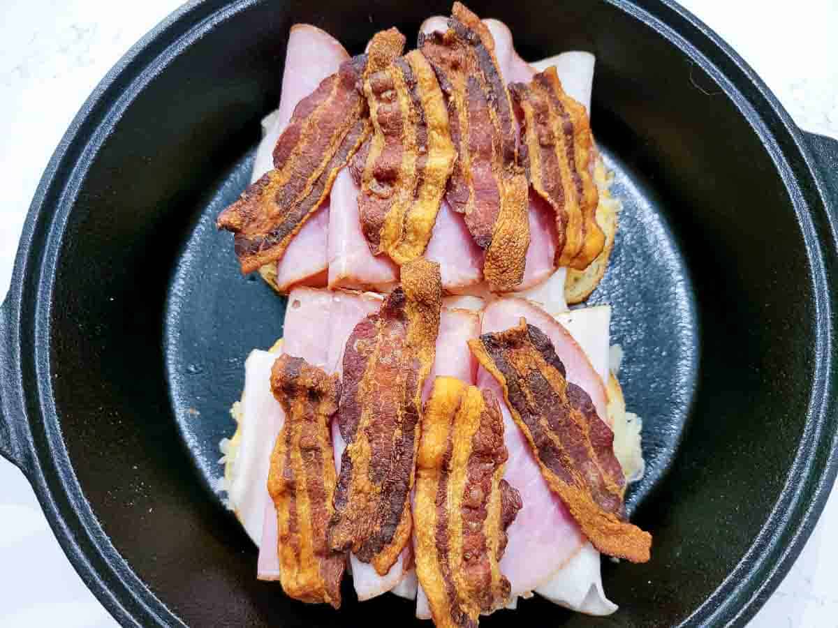 ham slices and cooked bacon slices added to two slices of rye bread in a cast iron skillet.